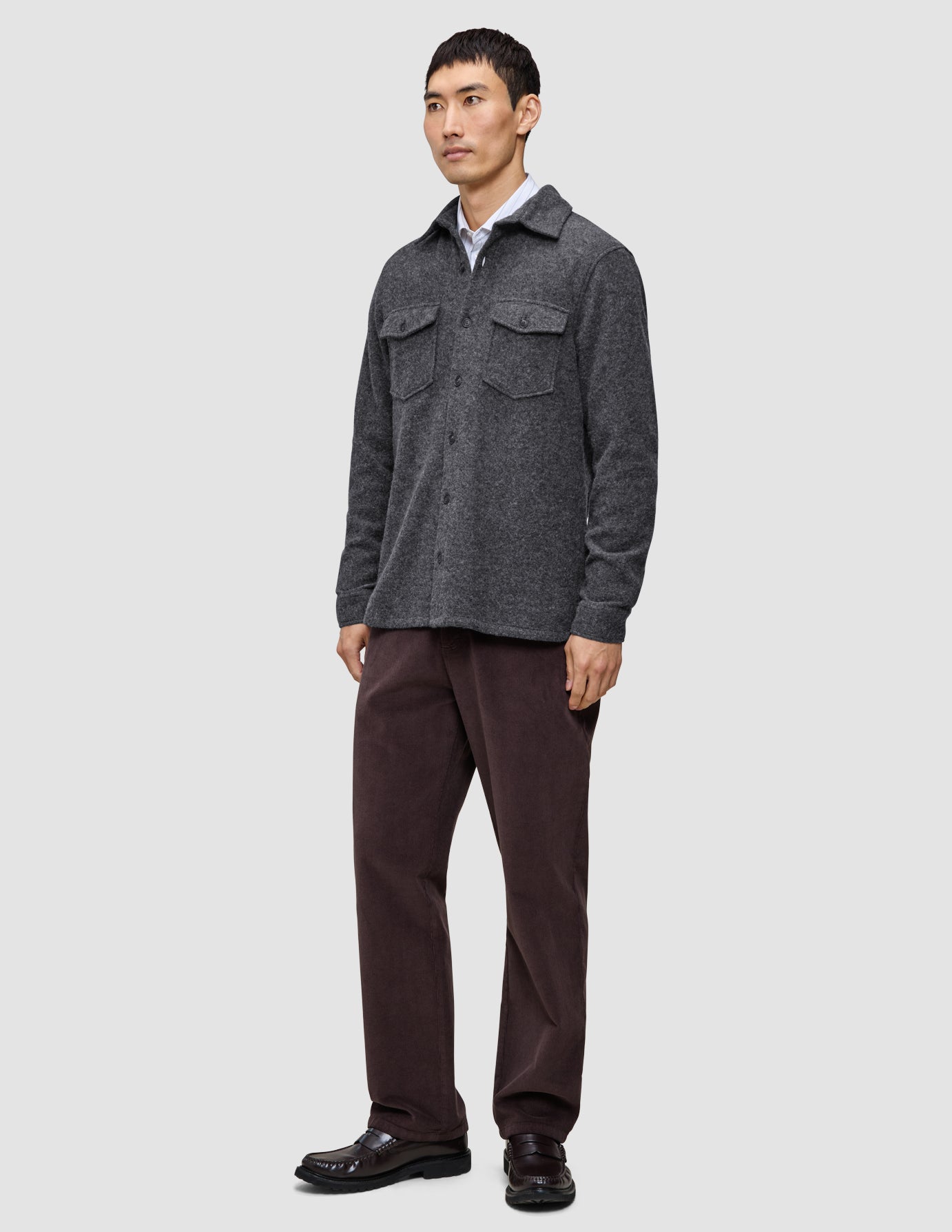 Ein Mann, der ein doppellagiges Woll-Overshirt in Regular-Passform und Dunkelgrau über einem weißen Hemd mit Kragen trägt, dazu eine dunkle Hose und braune Slipper in einem schlichten, skandinavischen Design.